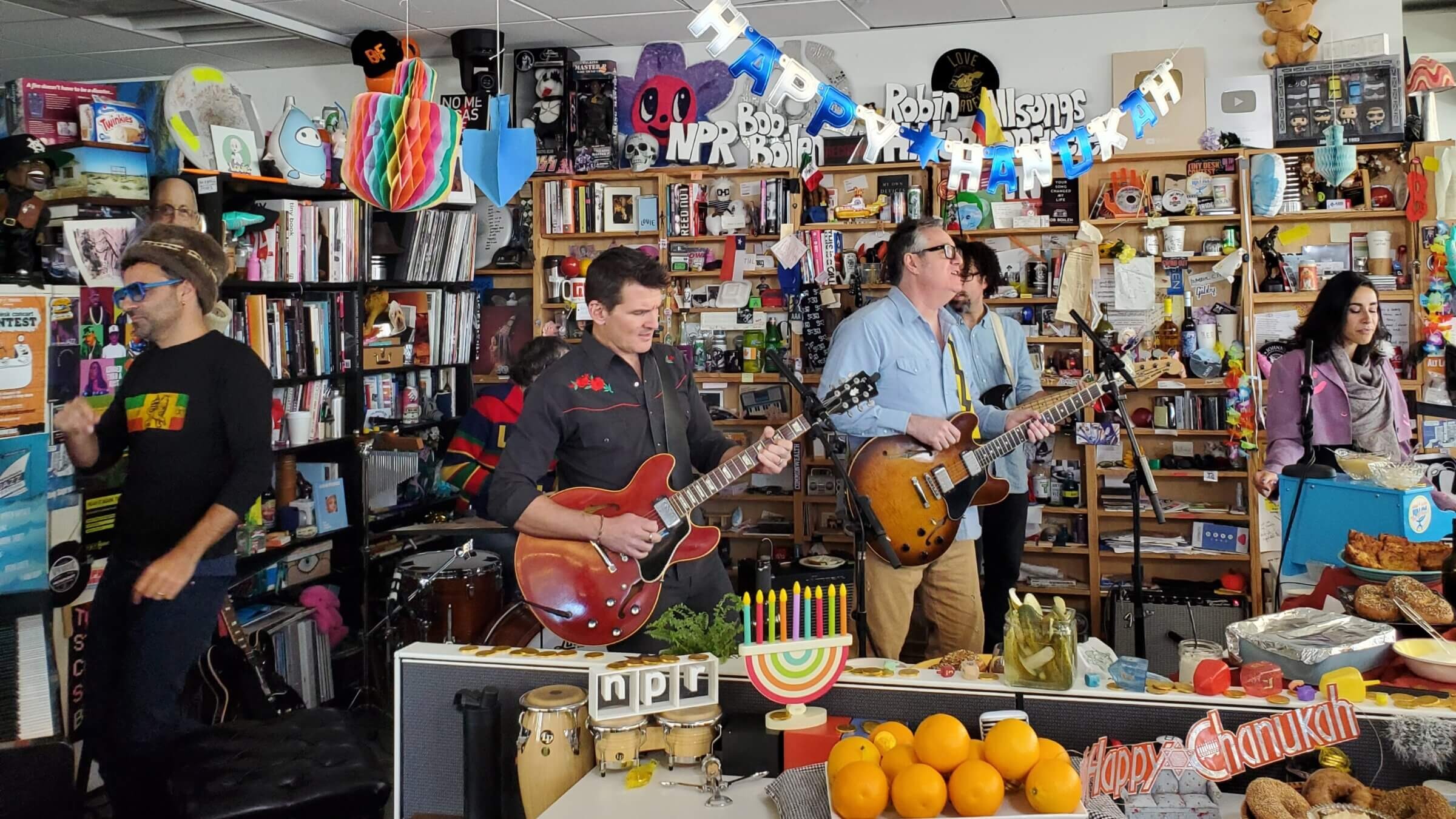 a male singer, two male guitarists, and a woman playing an instrument that is out of frame, rock out in the crowded, book-lined NPR Tiny Desk Concert venue