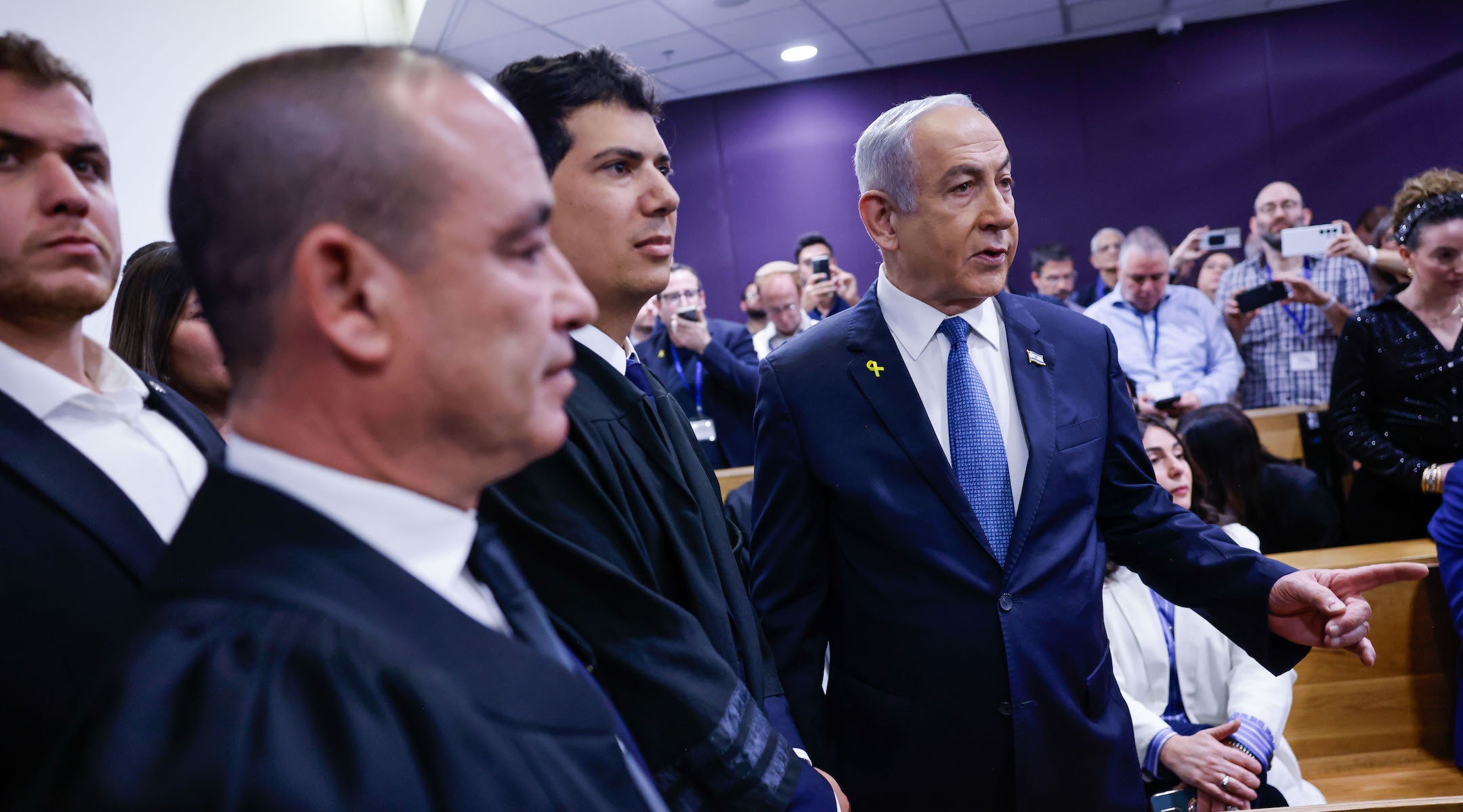 Netanyahu stands in a court room as two robed judges pass him
