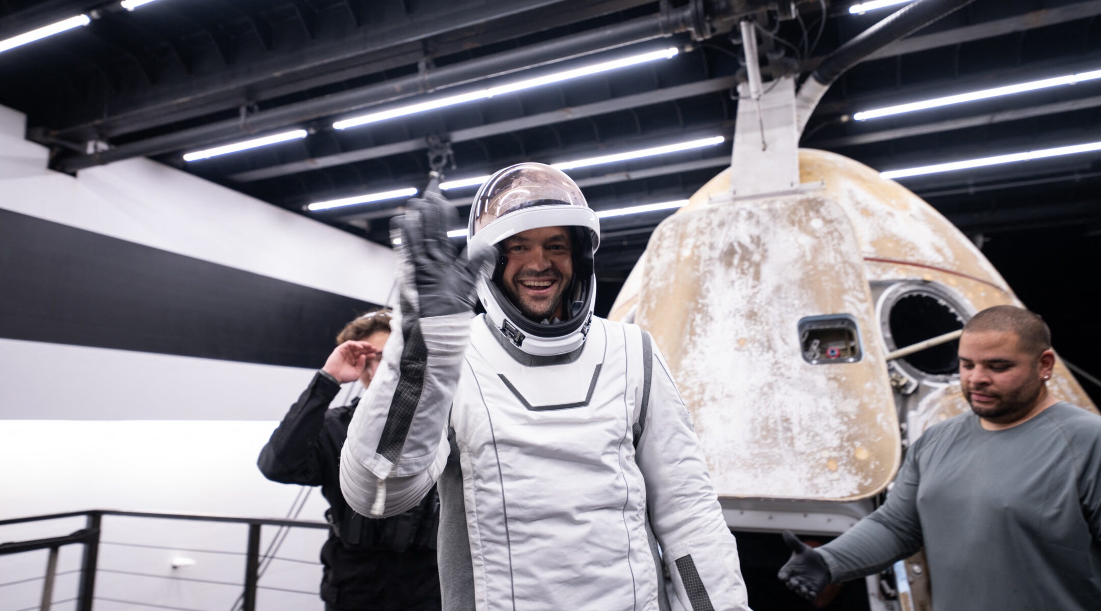 A man in a sleek spacesuit smiles and gives a thumbs-up signal. A small space capsule sits behind him.