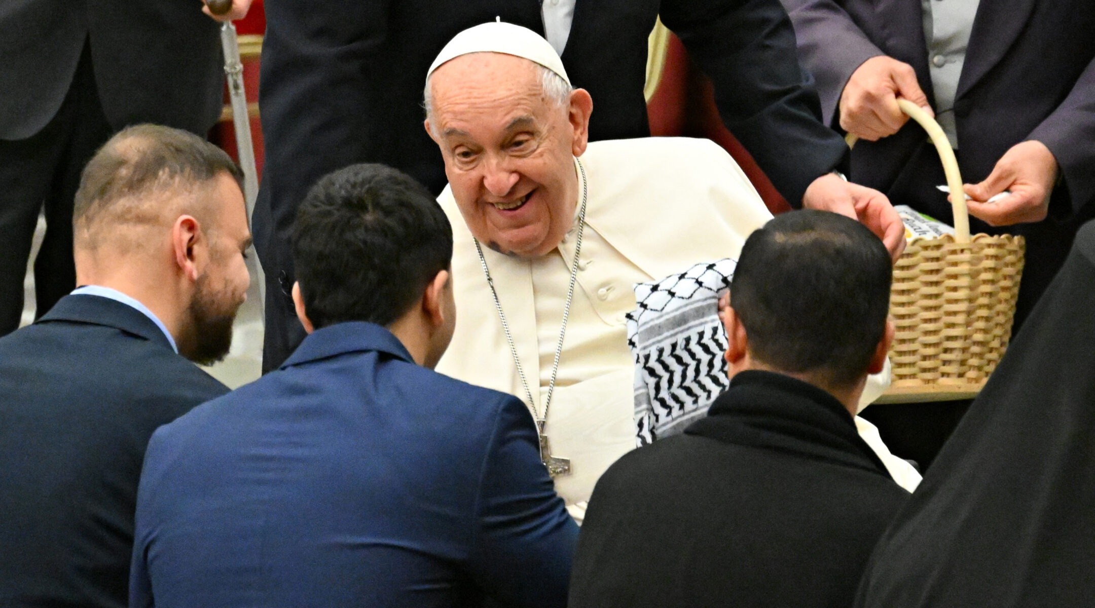 Pope Francis smiles as he holds a keffiyeh and talks with three men in suits who are seen from behind