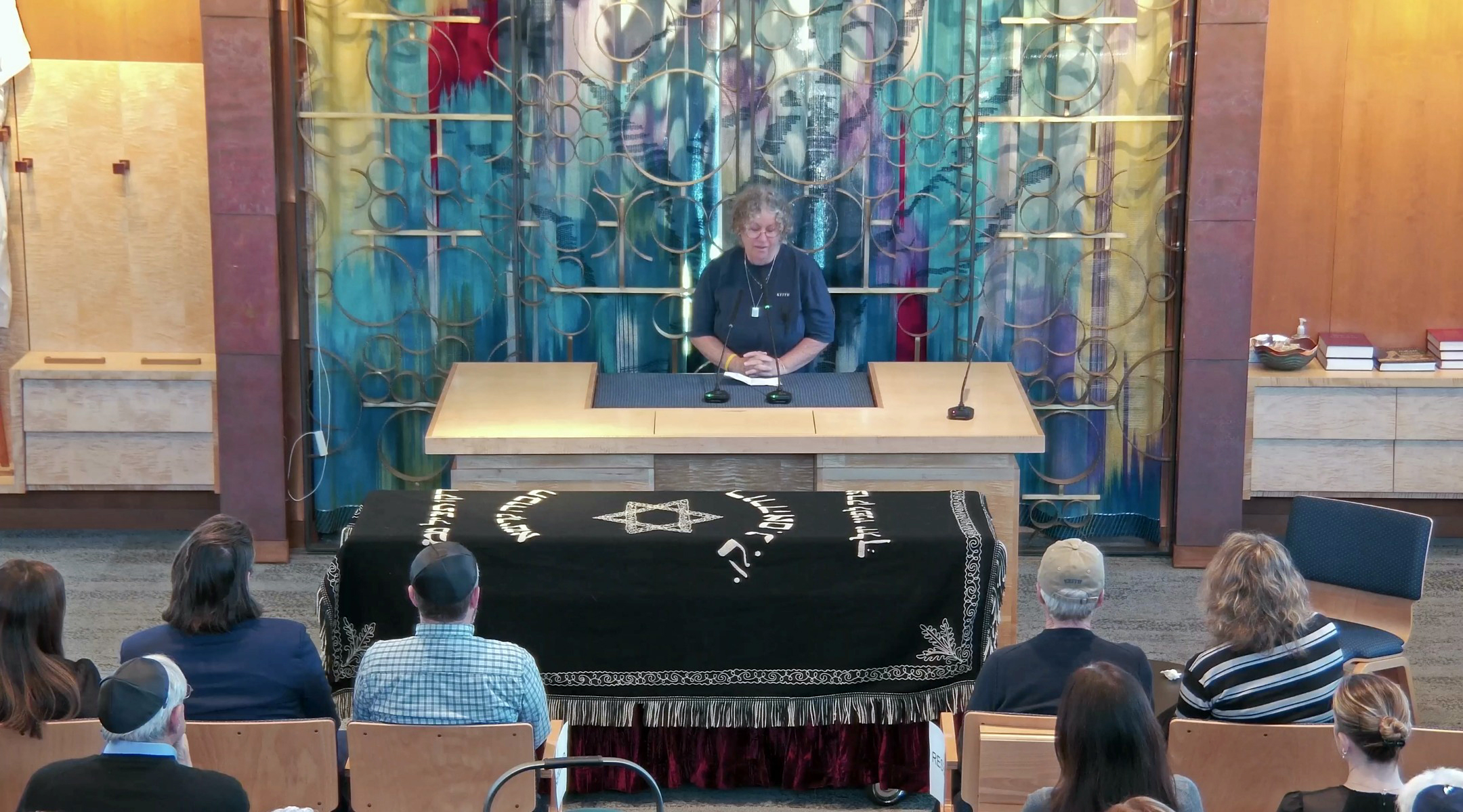 a woman speaks at the lectern on a wood-paneled synagogue sanctuary