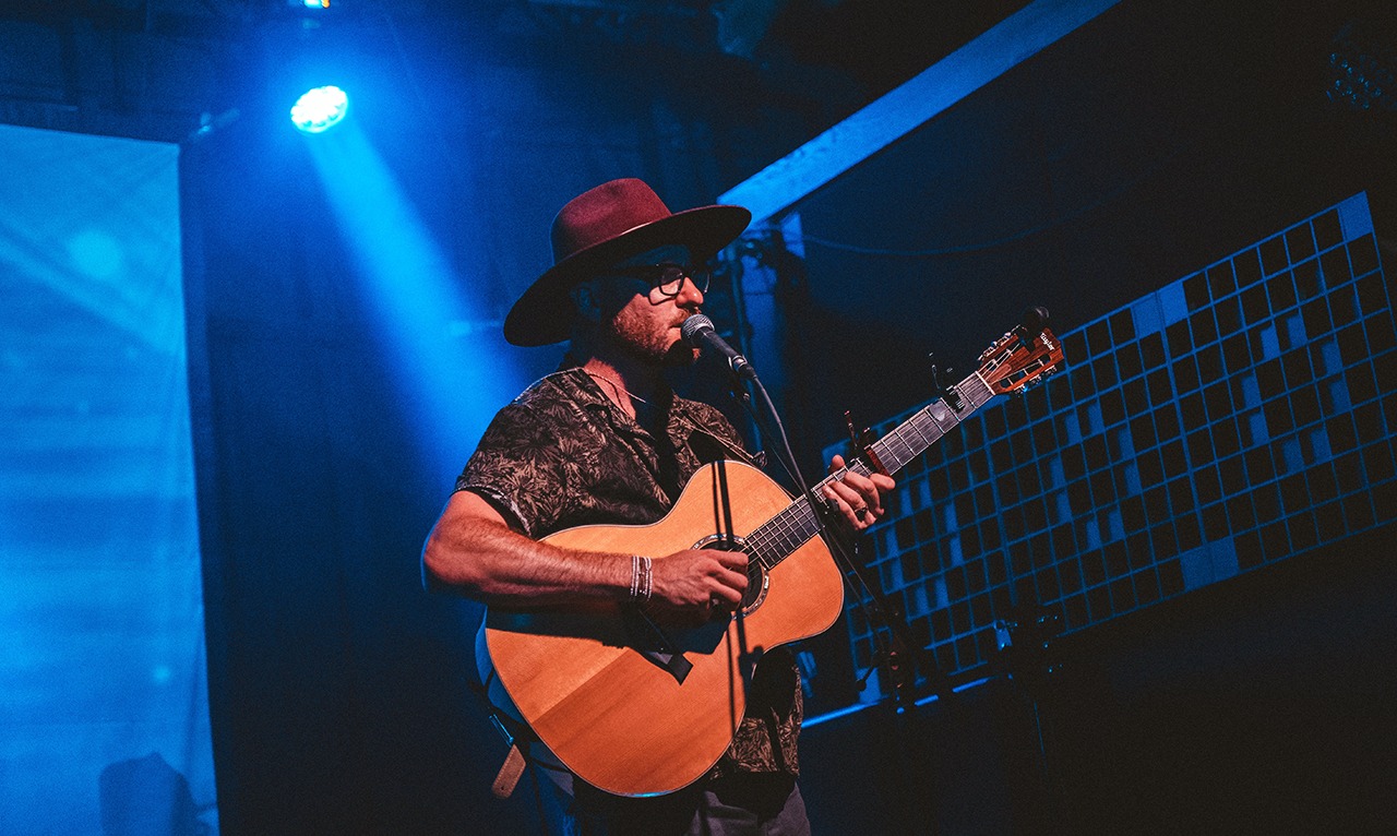 A man in a wide-brimmed hat plays guitar on stage while singing into a microphone