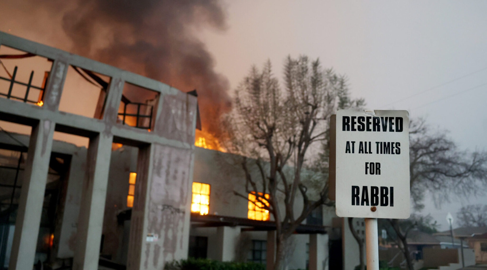 a sign in a parking lot reads "RESERVED AT ALL TIMES FOR RABBI" as a synagogue building burns in the background