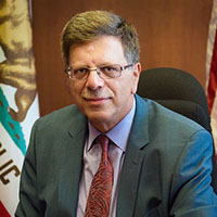 a posed headshot of a man in a suit with a mustache. a california flag hangs behind him.