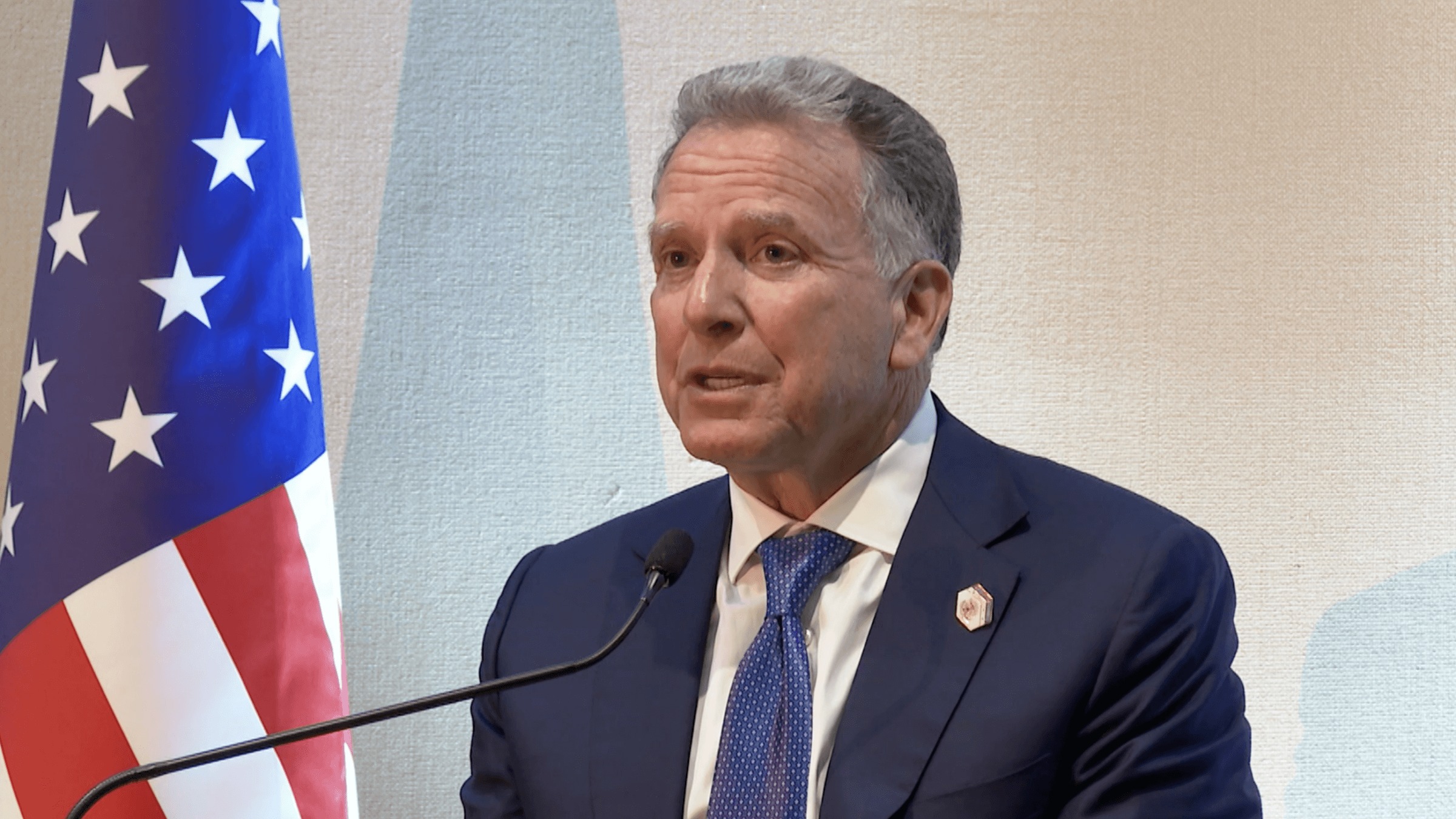 An older man in a suit speaks with an American flag behind him