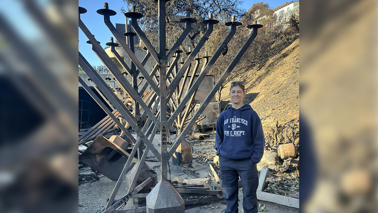 A woman in a blue hoodie stands in front of a tall, fire-marked menorah outside