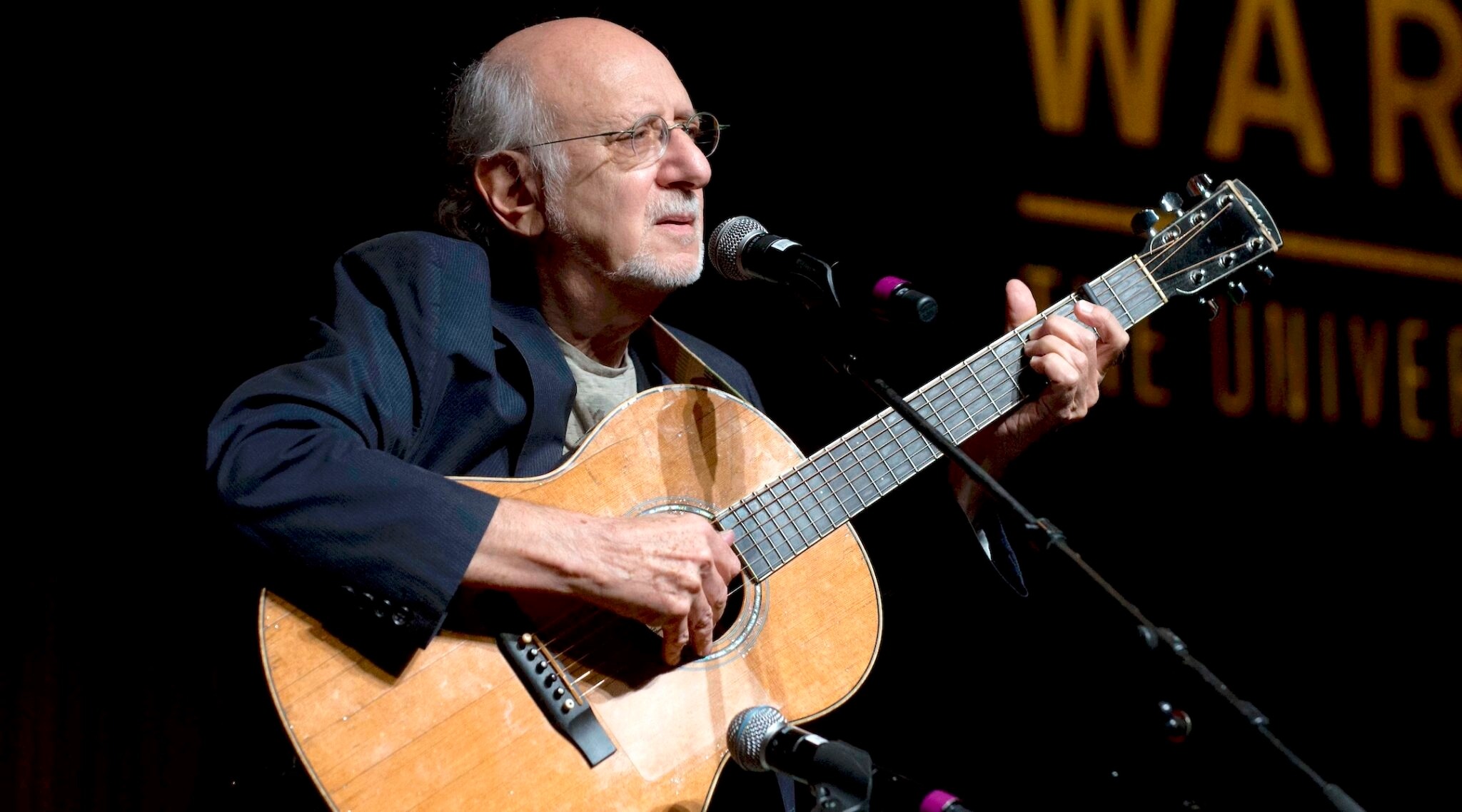 An older bald man plays guitar and sings into a microphone
