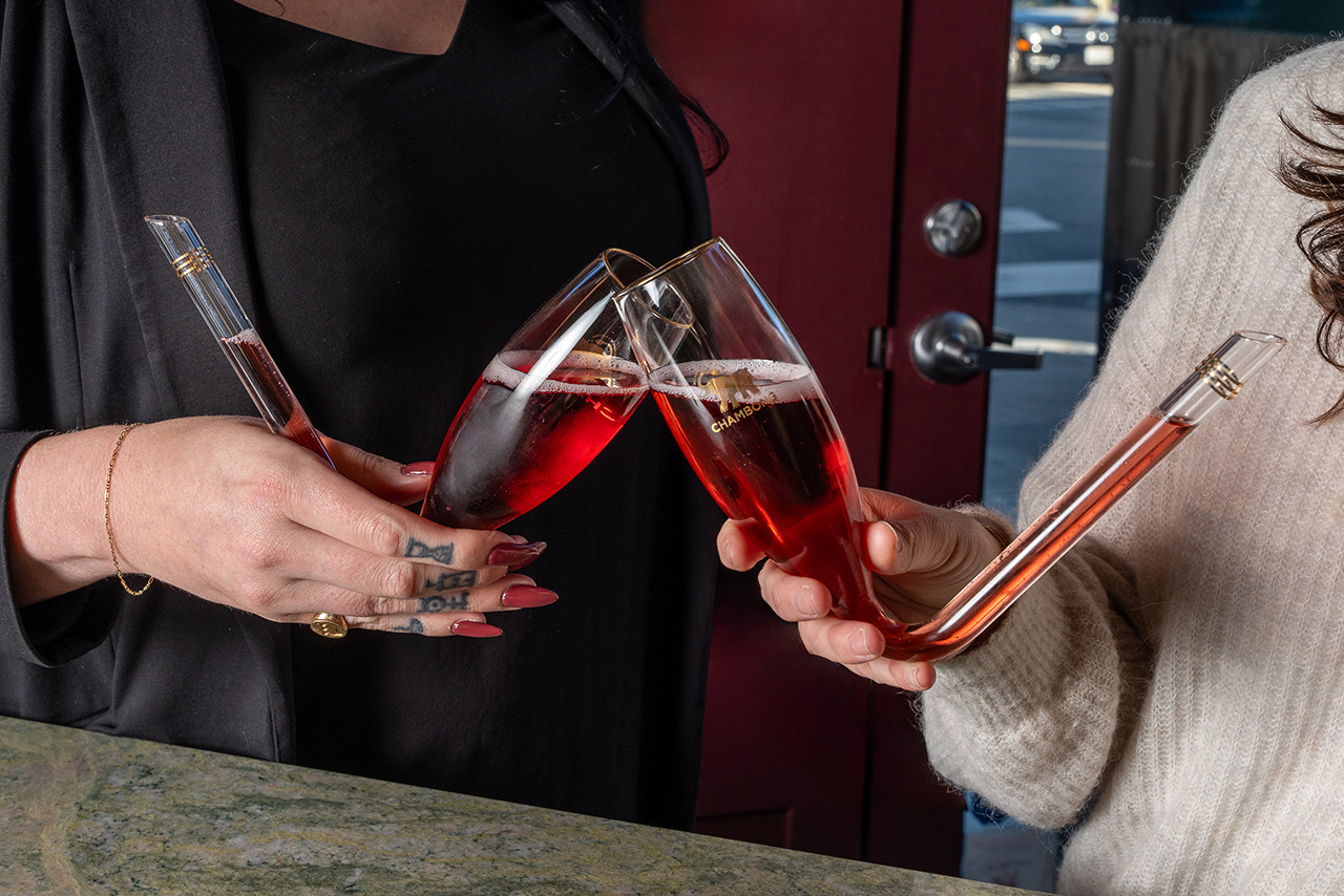 a close up of two people's hands holding "chambongs" — which resemble champagne flute glasses, but are held at an angle with the stem curving away from the glass as a straw