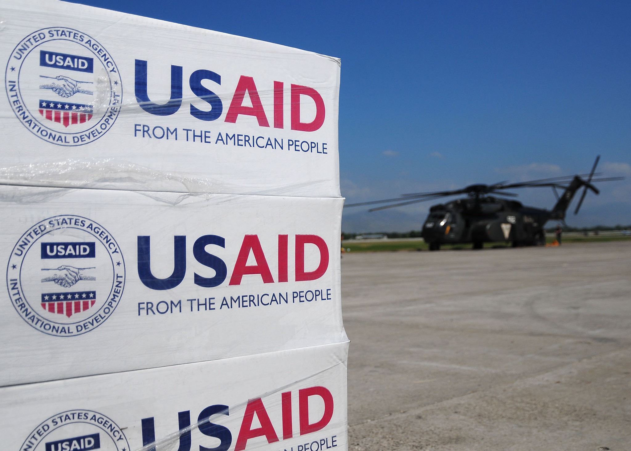 In the foreground, there is a stack of boxes that said USAID on them. In the background, a military helicopter sits on the tarmac.