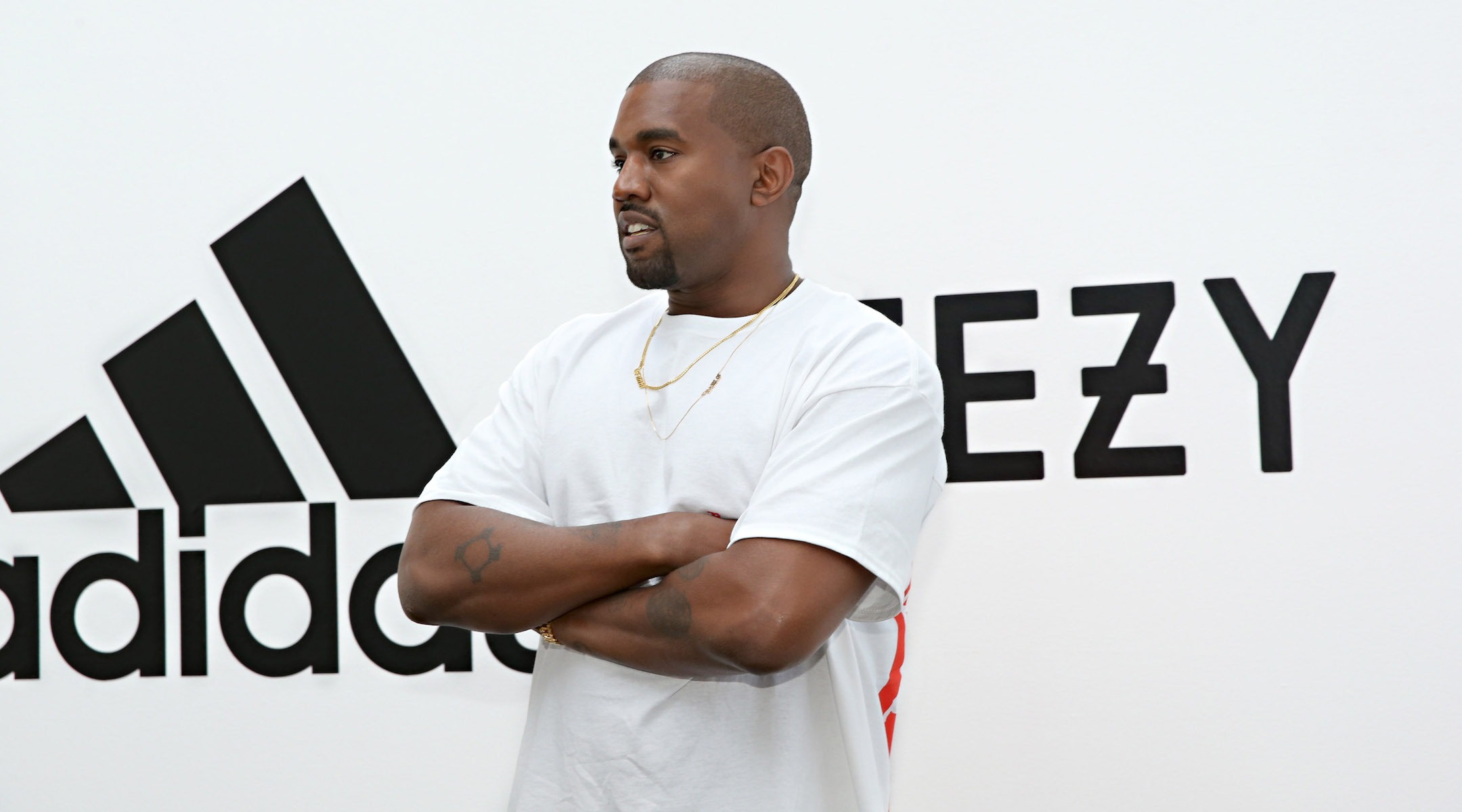 Kanye stands with his arms crossed in front of a sign with the Adidas and Yeezy logos