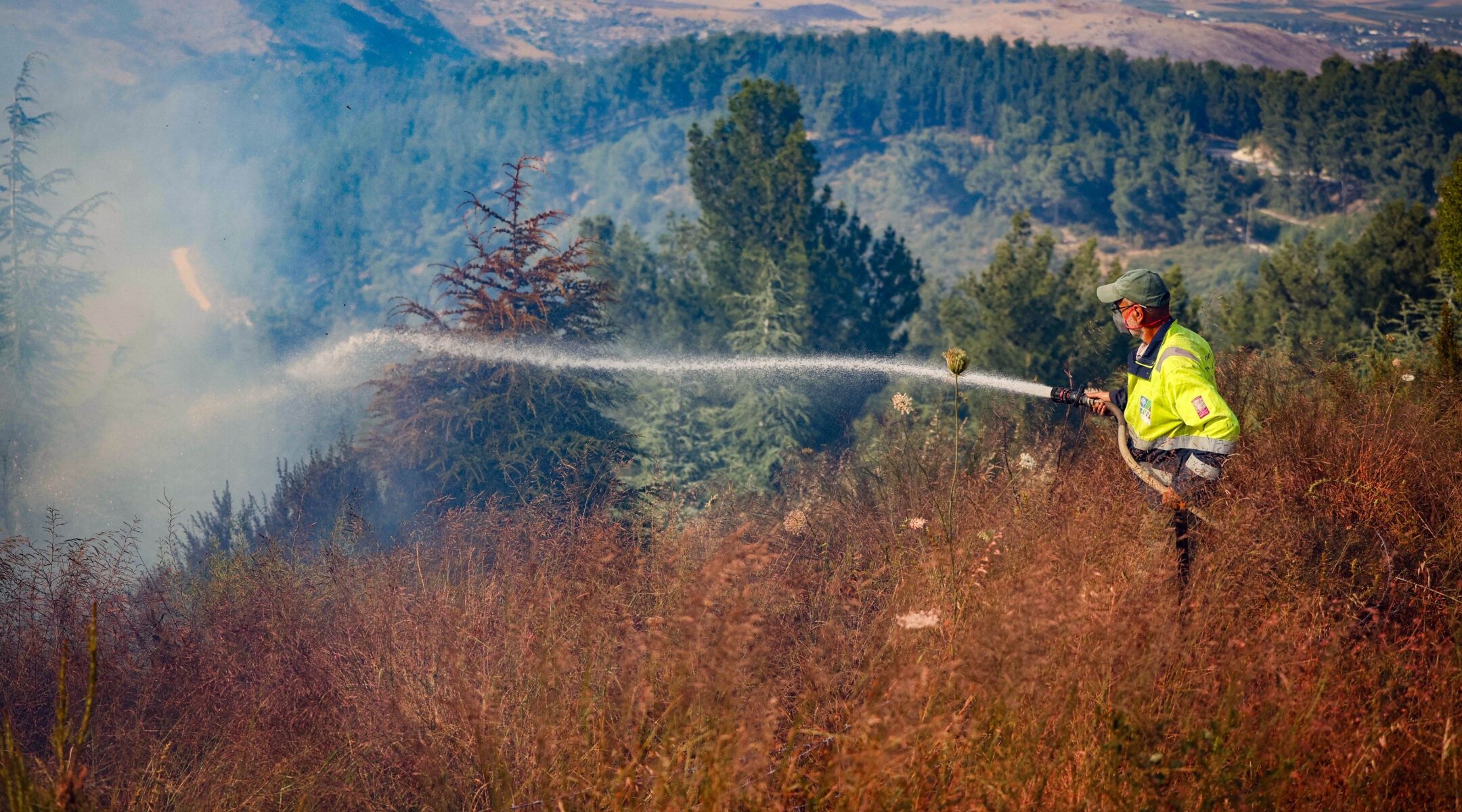A firefighter stands on a high ridge, spraying brush with a firehose