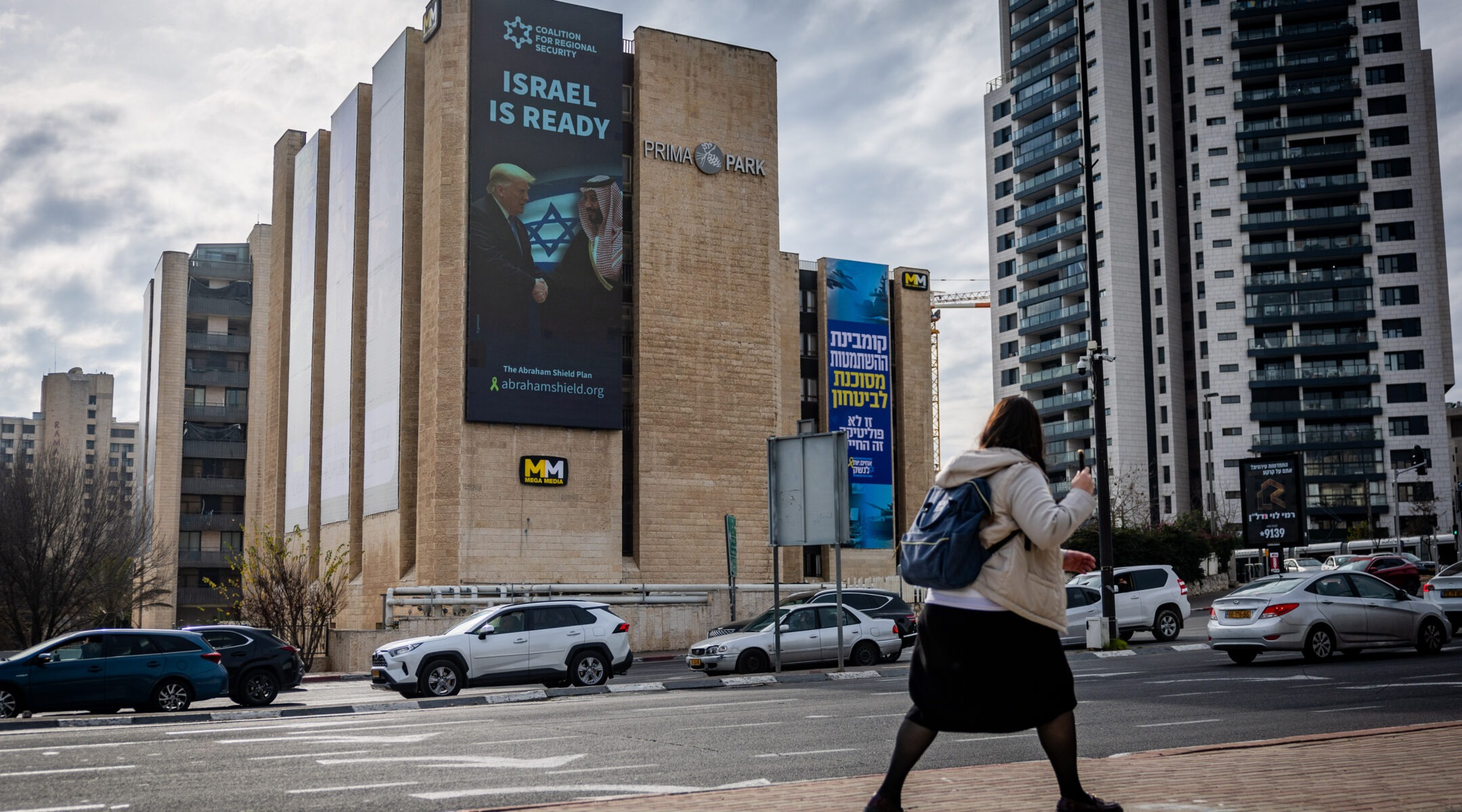 A woman walks past a building with a large image of Trump and Mohammed bin Salman shaking hands. The text on the image reads "Israel is ready."
