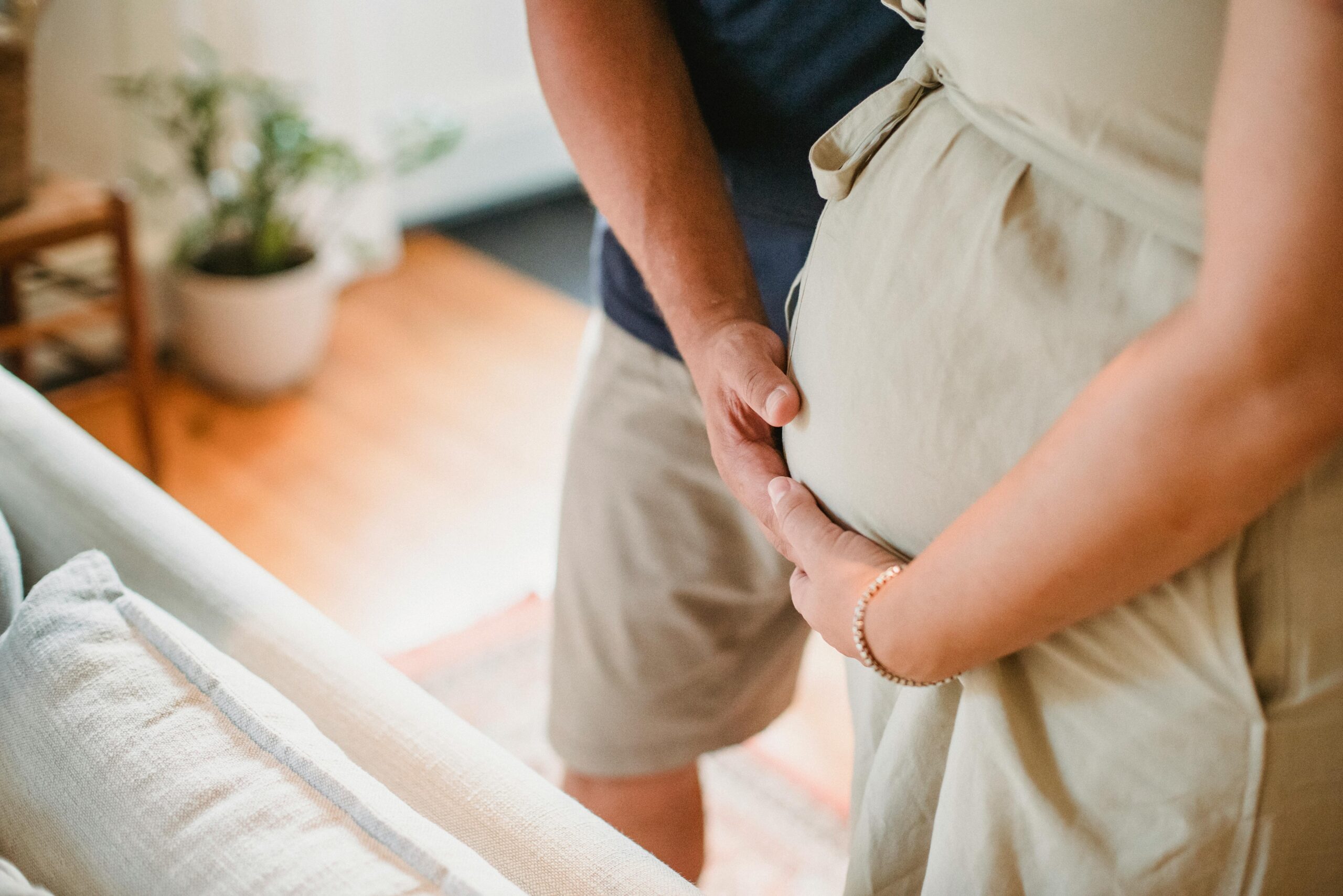 Stock photo shows a closeup of a pregnant woman's belly under a beige dress. her arm and the arm of a man standing next to her cradle her belly.