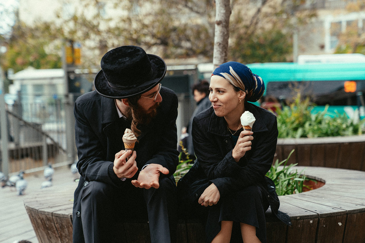 A Haredi man in a black hat and black coat sits on a park bench eating ice cream with a Haredi woman with covered hair