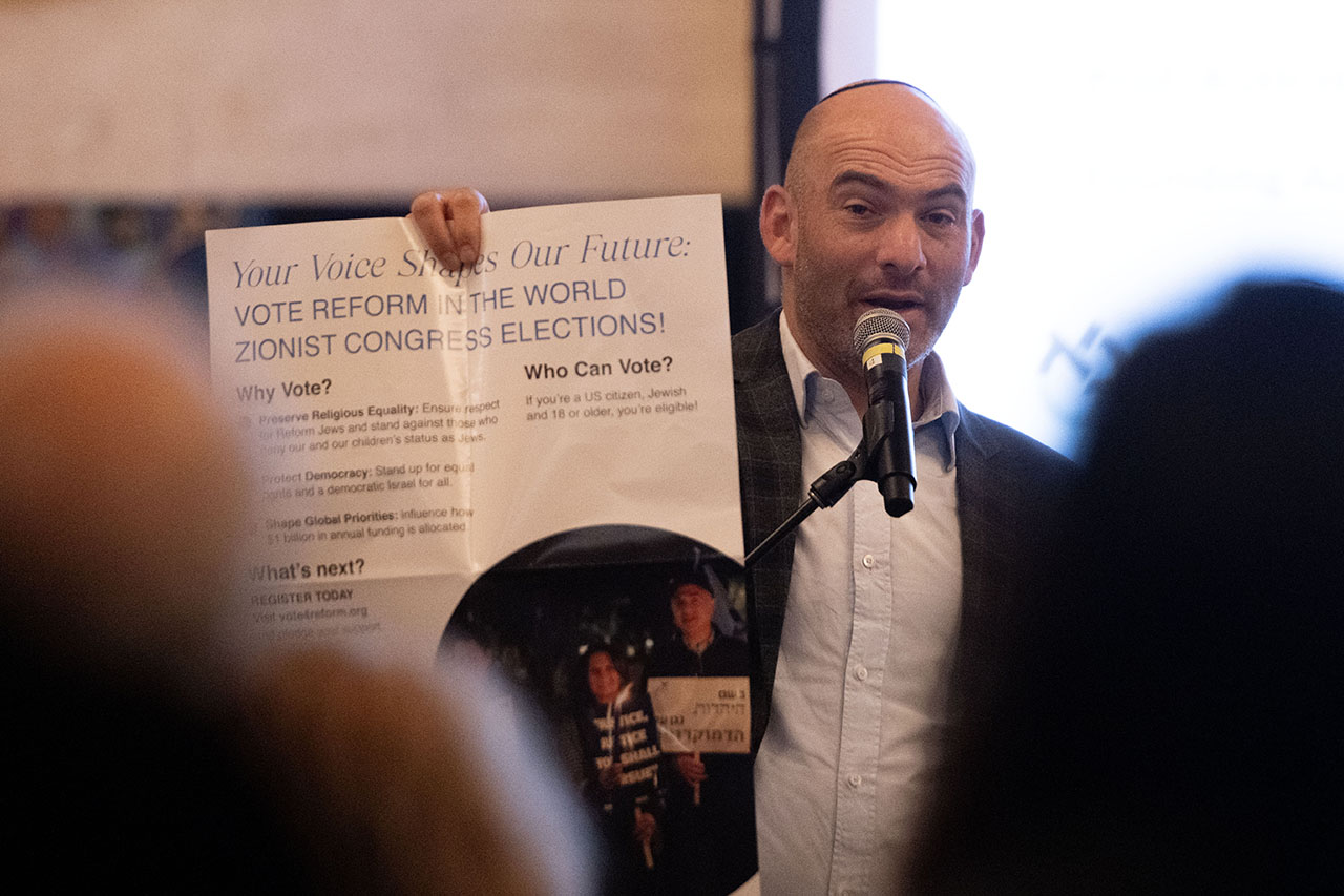 a bald middle-aged man in a suit holds up a poster/brochure titled "Your Voice Shapes Our Future" as he speaks to an indoor audience