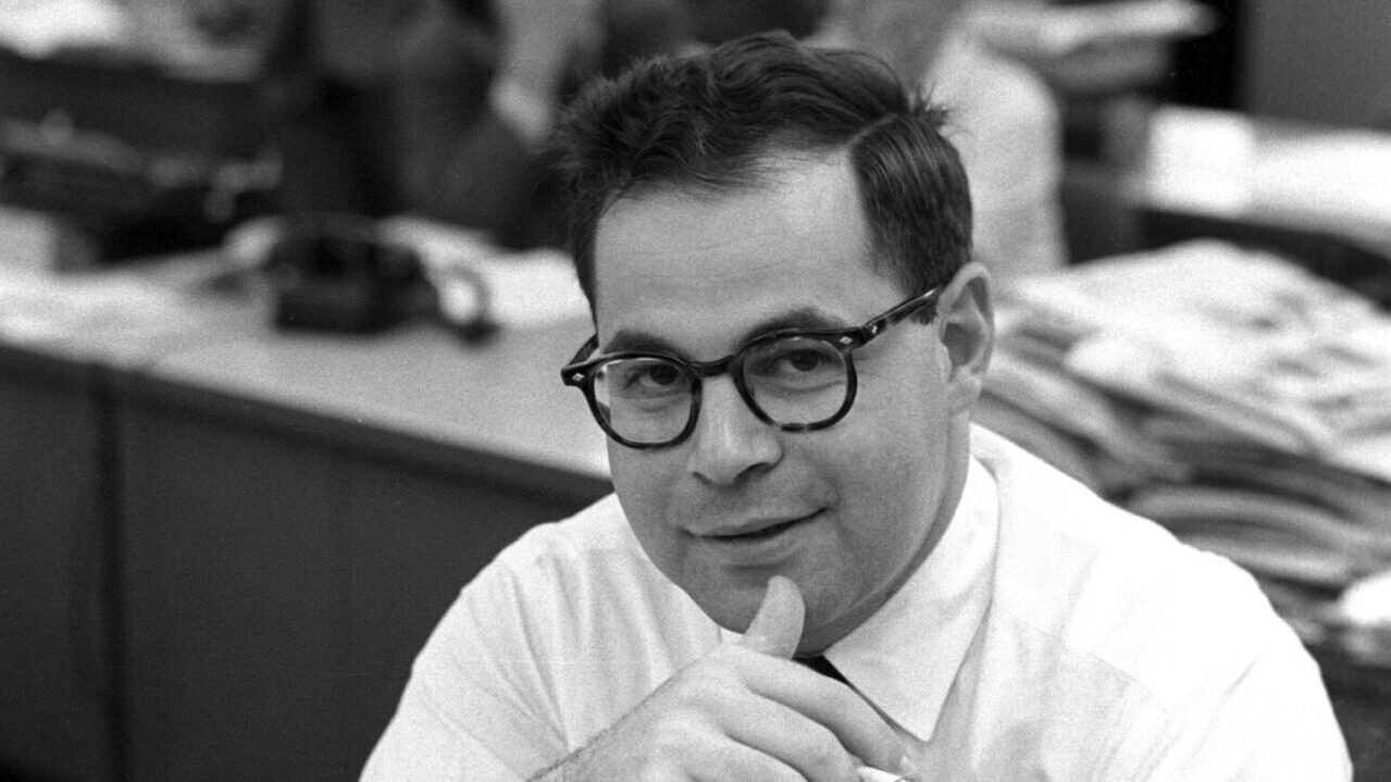 A black and white photo of a man in glasses sitting at a desk with a slight grin