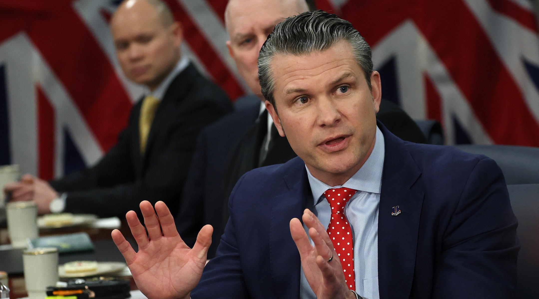 A white man in a suit gestures while speaking in a meeting