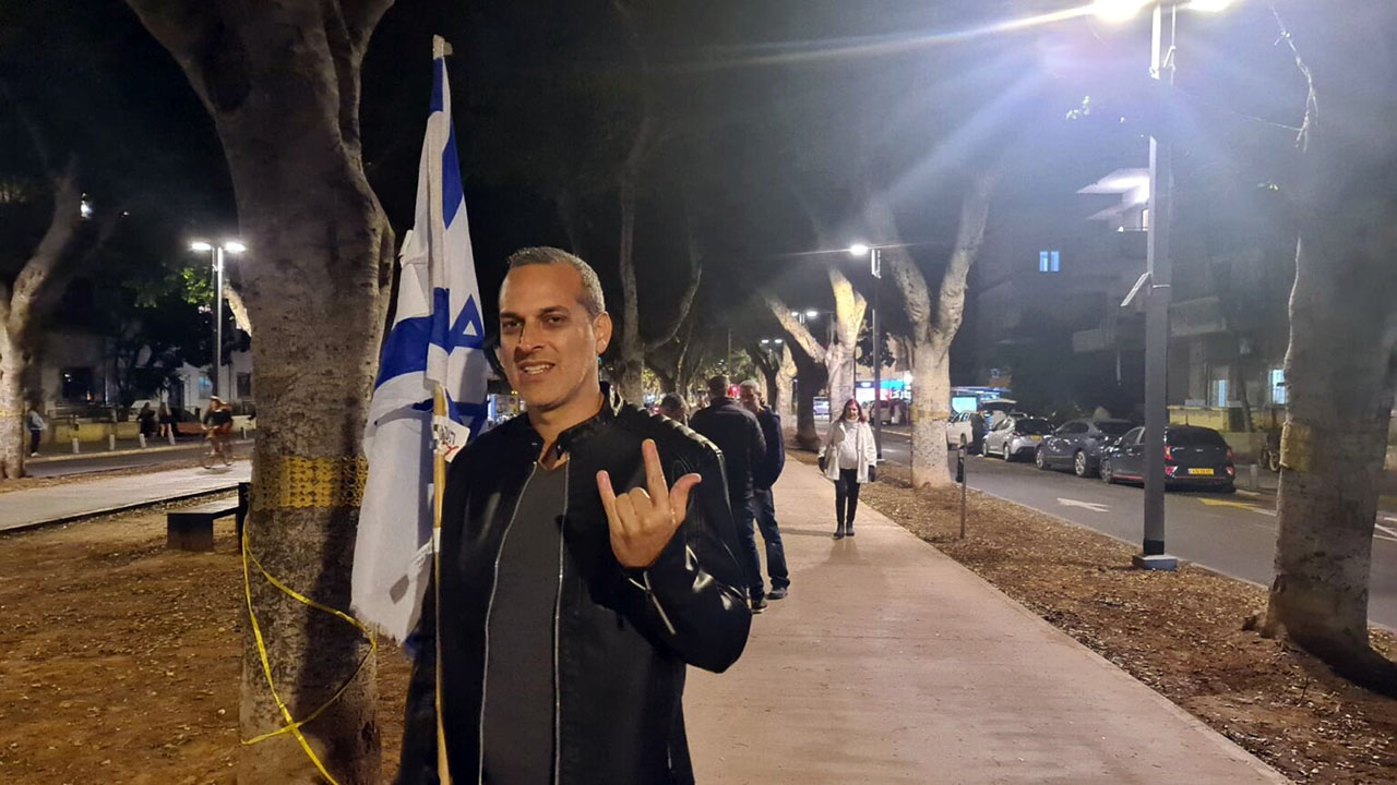 A man in a leather jacket stands outside at night with an Israeli flag behind him