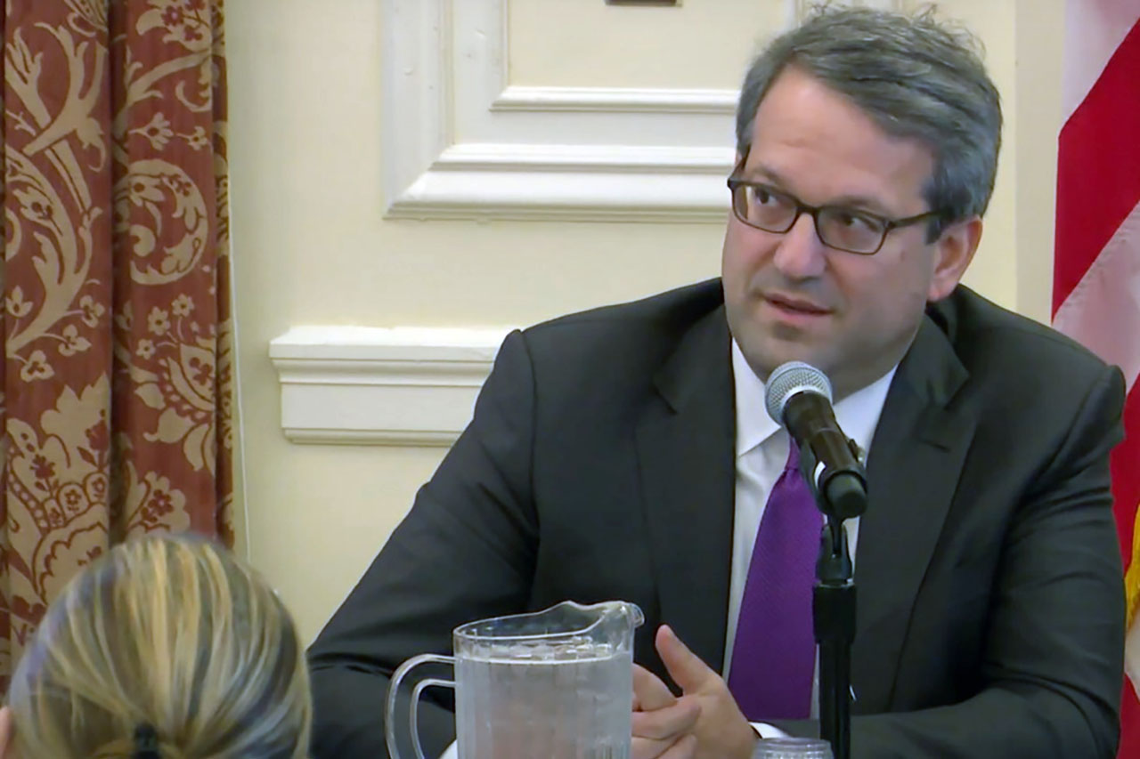A middle aged white man in a suit sits speaking into a microphone