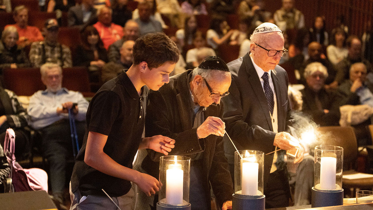 Three men, one very young and two very old, light candles in front of a packed auditorium