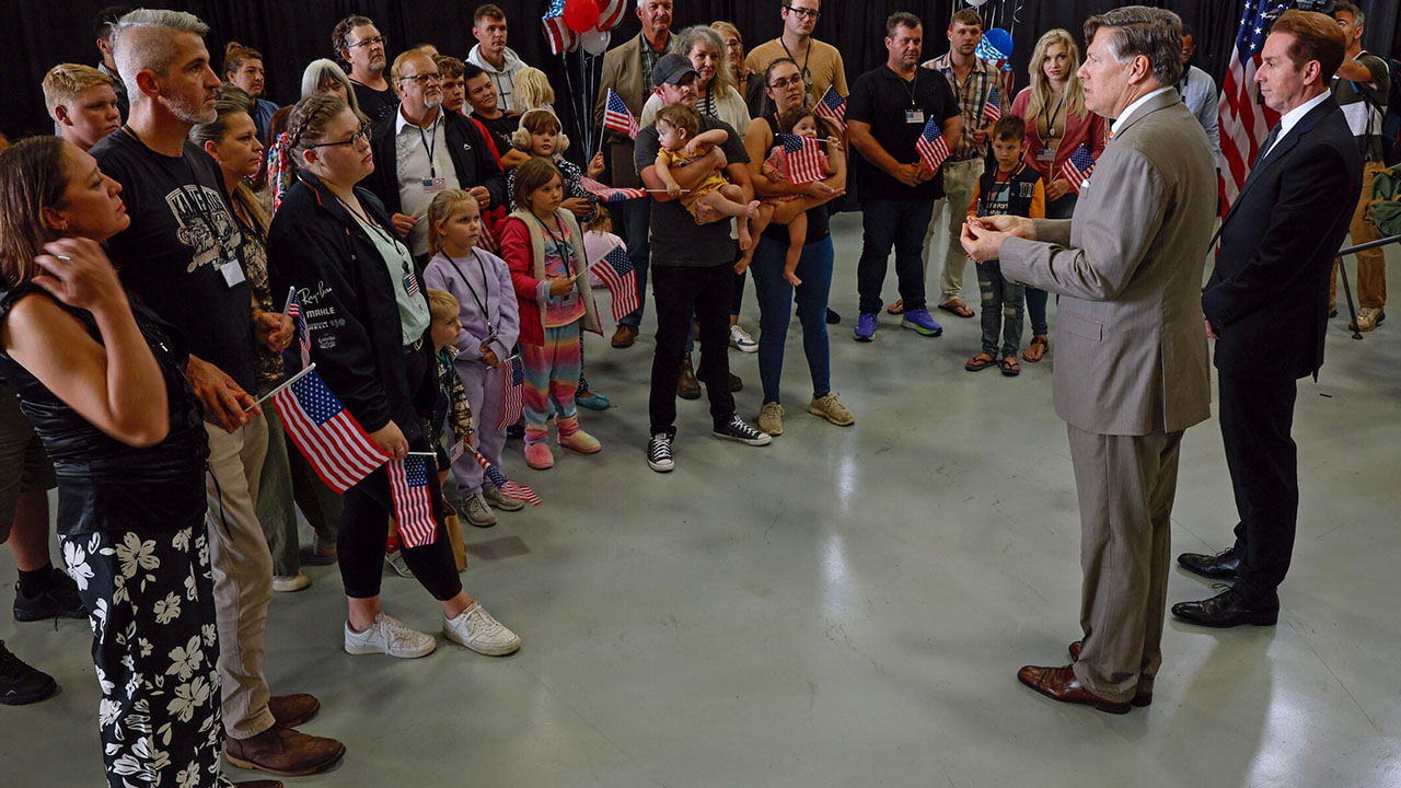 Newly arrived South Africans listen to U.S. Deputy Secretary of State Christopher Landau and Homeland Security Deputy Secretary Troy Edgar (right) deliver welcome statements in a hangar near Washington Dulles International Airport on May 12, 2025. (Chip Somodevilla/Getty Images)