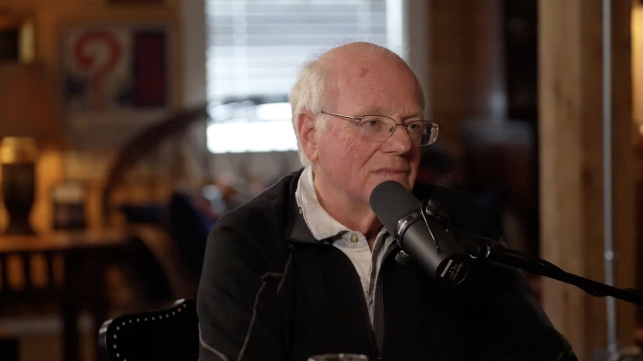 And older bald man in glasses sits speaking into a microphone