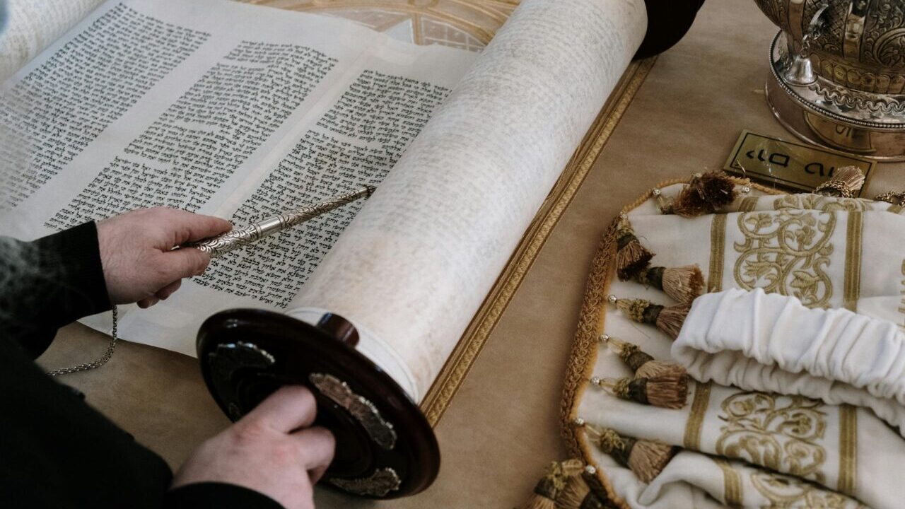 Person reading a Torah scroll using a yad in a traditional synagogue setting.