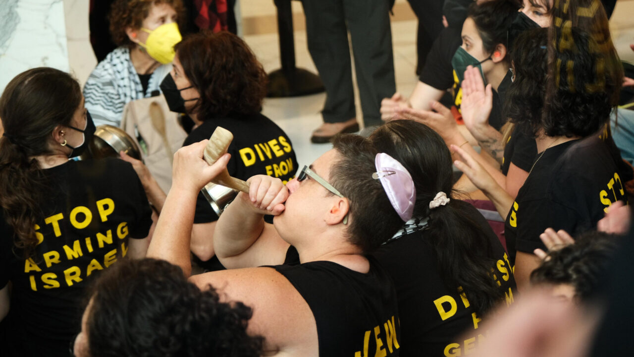 Person with kippah blows a small shofar while wearing a Jewish Voice for Peace shirt