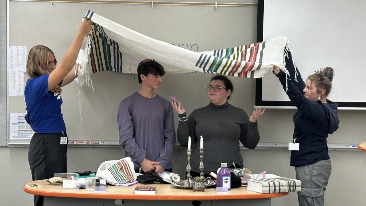 Two teens stand under a tallit while two others hold it.