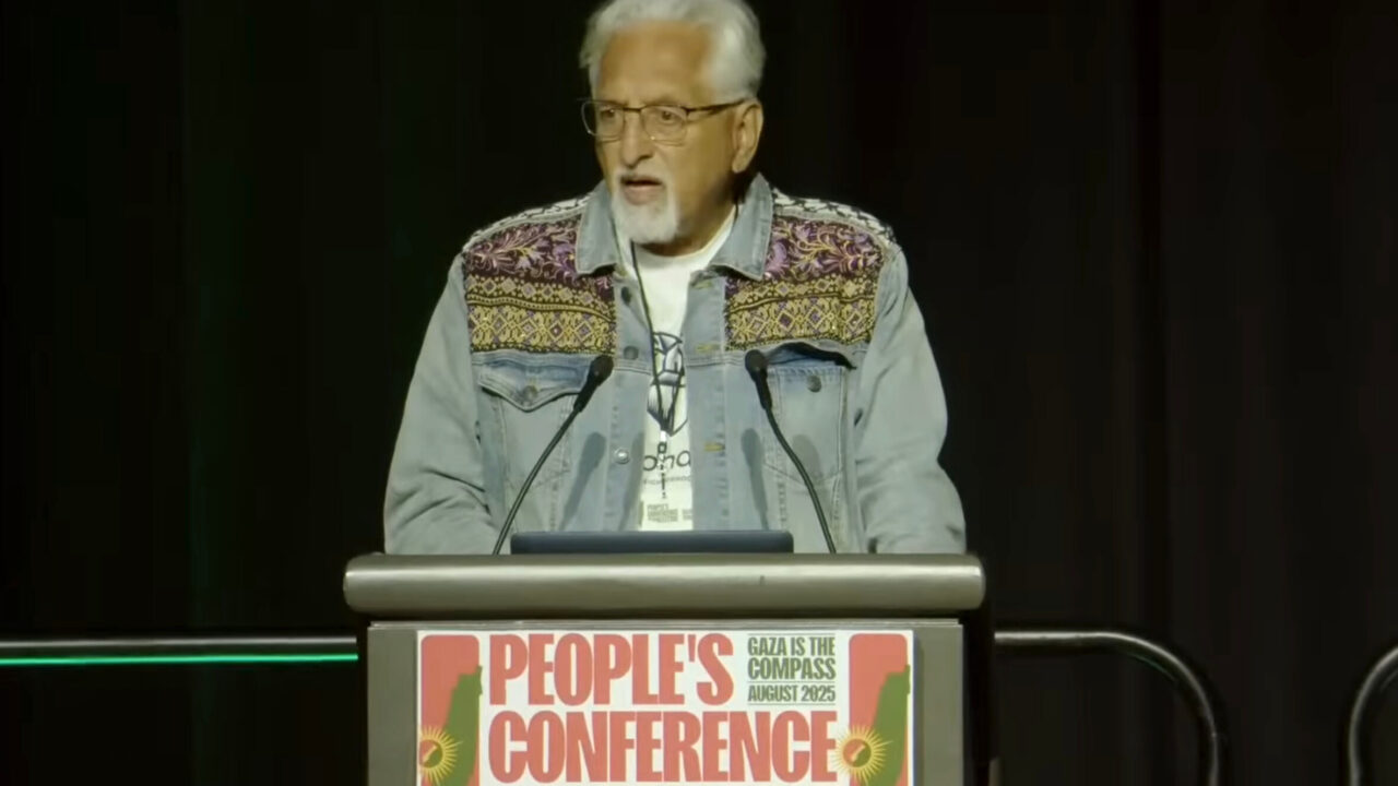 man with white hair, classes and decorative jean jacket speaks at podium with sign reading 'People's Conference" + "Gaza Is the Compass"