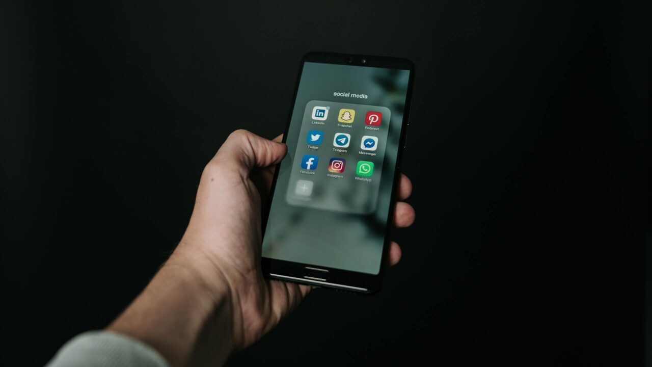 Close-up of a hand holding a smartphone displaying various social media app icons on a dark background.