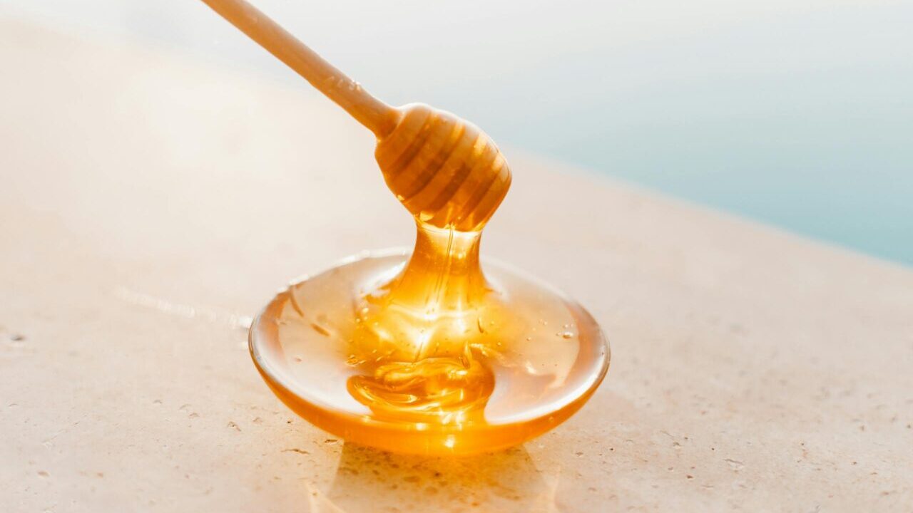 Golden honey flowing from a wooden dipper onto a glass plate, captured in warm daylight.