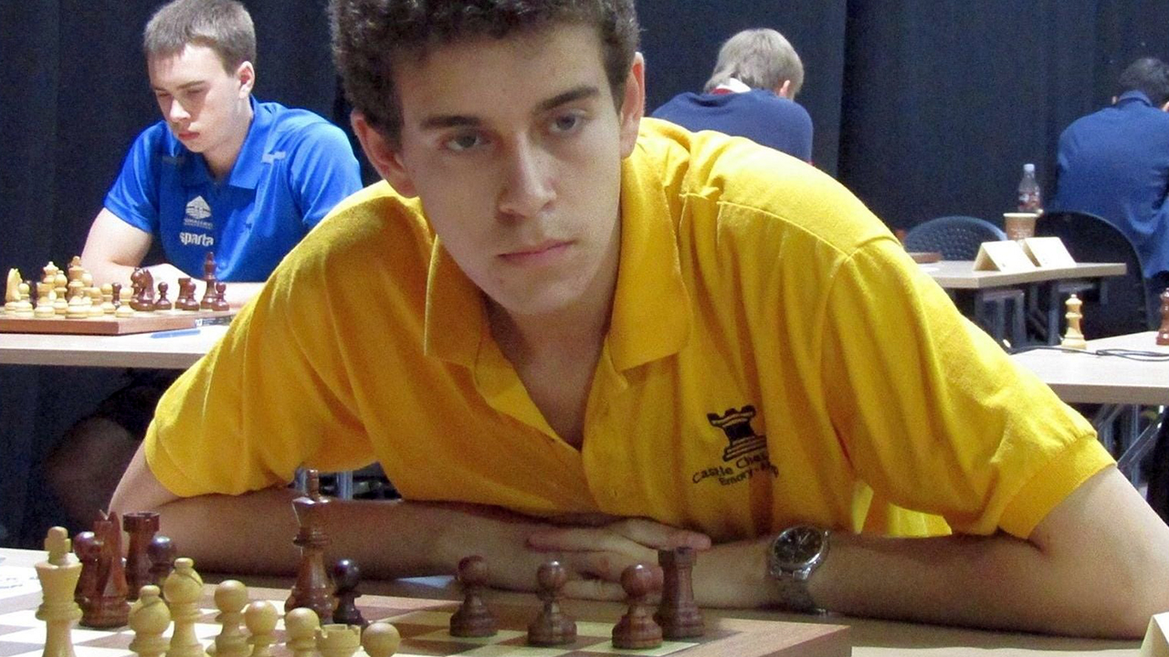Naroditsky, seated, leans forward and gazes at a chess board
