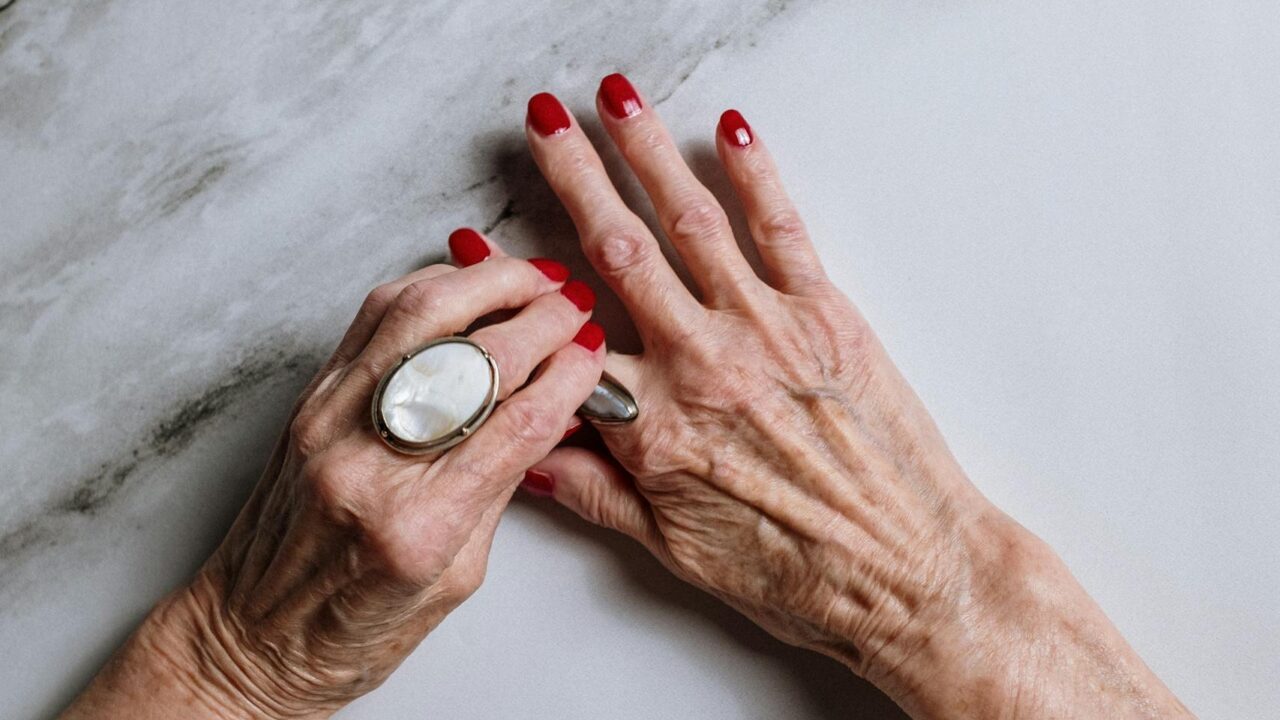 Beautifully manicured senior hands with red nails and jewelry on a marble surface.
