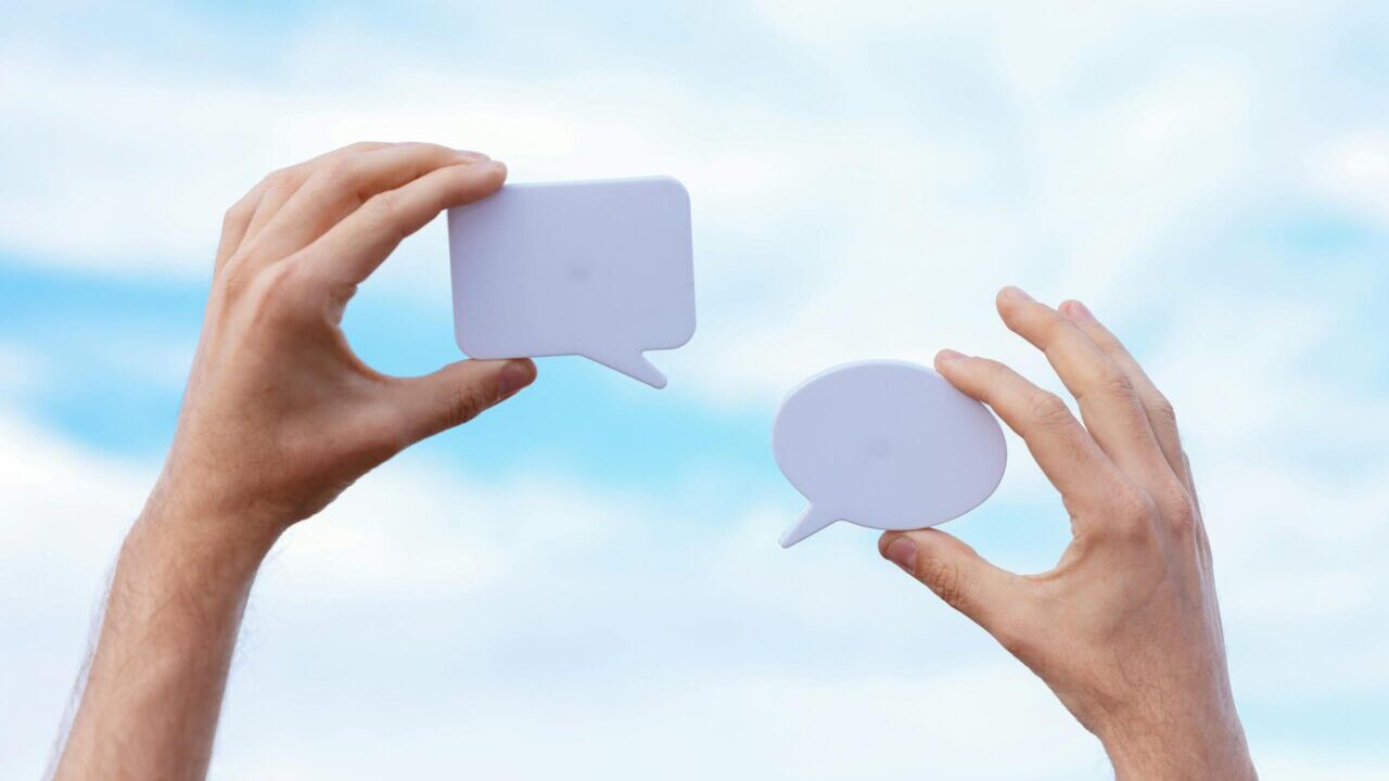 Close-up of hands holding speech bubbles, symbolizing communication and dialogue against a blue sky.